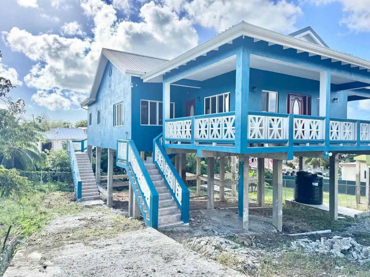 Newly constructed teal and white two-storey house elevated on concrete pillars with a large covered front patio and decorative white balustrade railings, Saint Lucia