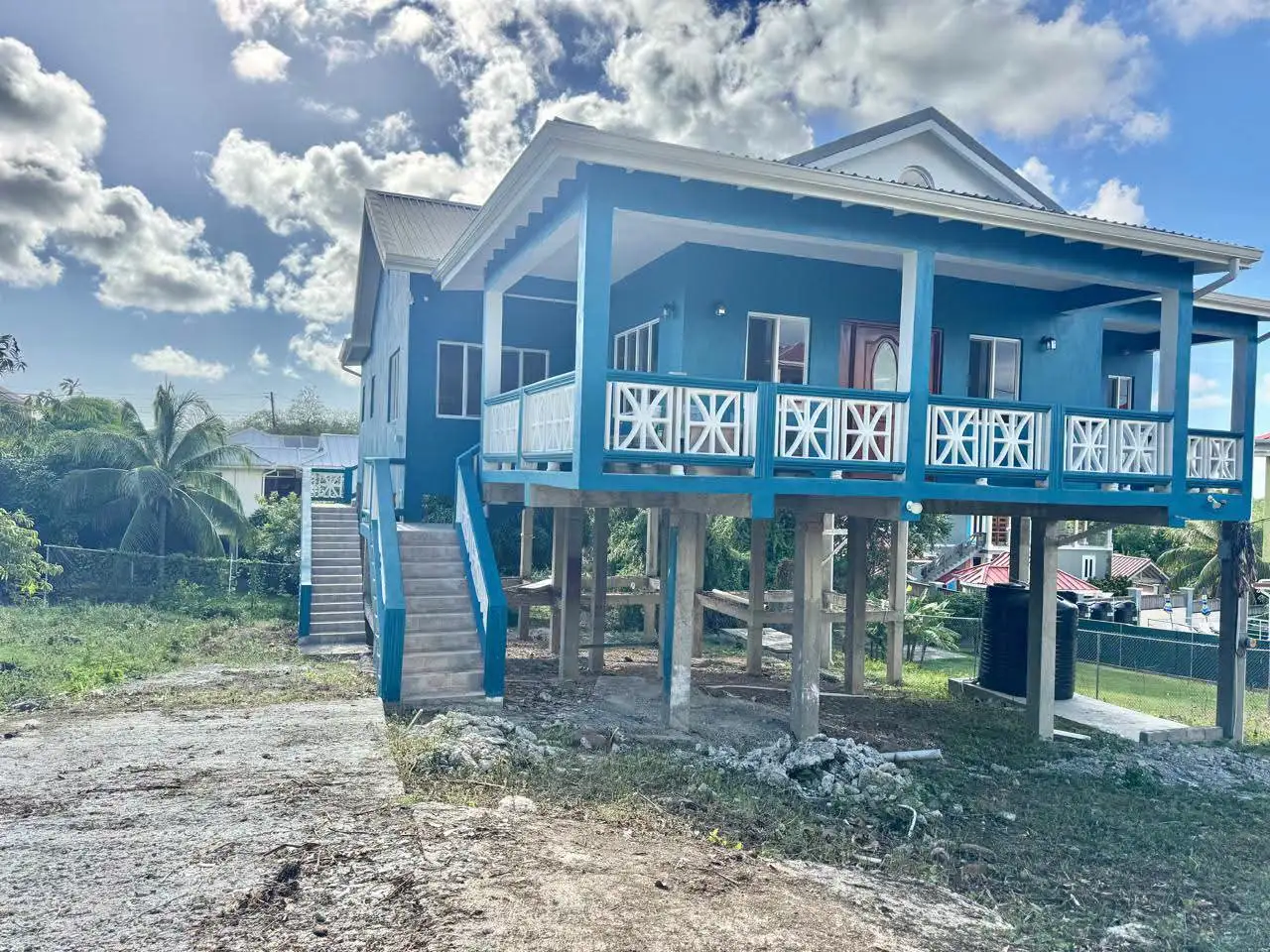 Second angle of newly constructed teal and white elevated house showing spacious covered outdoor patio area and ground floor space beneath the structure, Saint Lucia