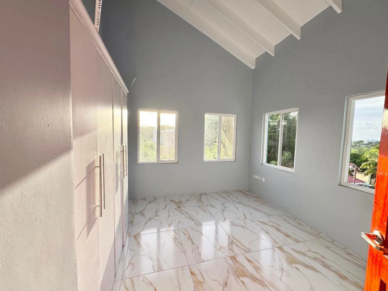 Spacious open-plan interior living area with high vaulted ceiling, cream walls, large white marble-design porcelain floor tiles and built-in wardrobe, Saint Lucia
