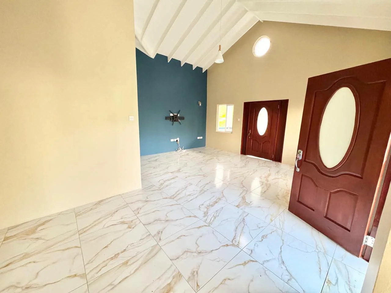 Open-plan living room with high pitched ceiling, warm cream and teal accent wall, polished marble-design floor tiles and solid mahogany front doors with oval glass inserts, Saint Lucia