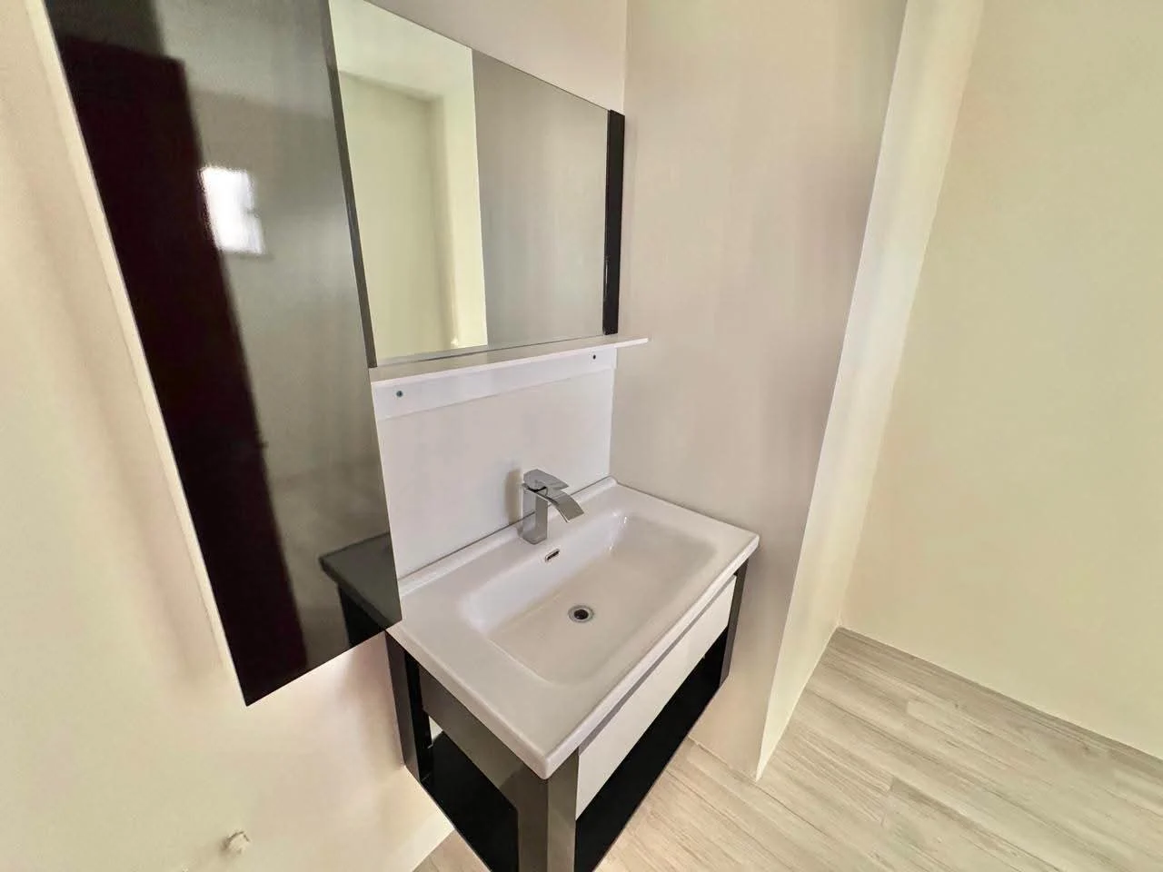 Modern bathroom vanity with white ceramic basin sink, chrome mixer tap, wall-mounted rectangular mirror with dark trim surround, set against neutral cream walls and wood-look flooring, Saint Lucia