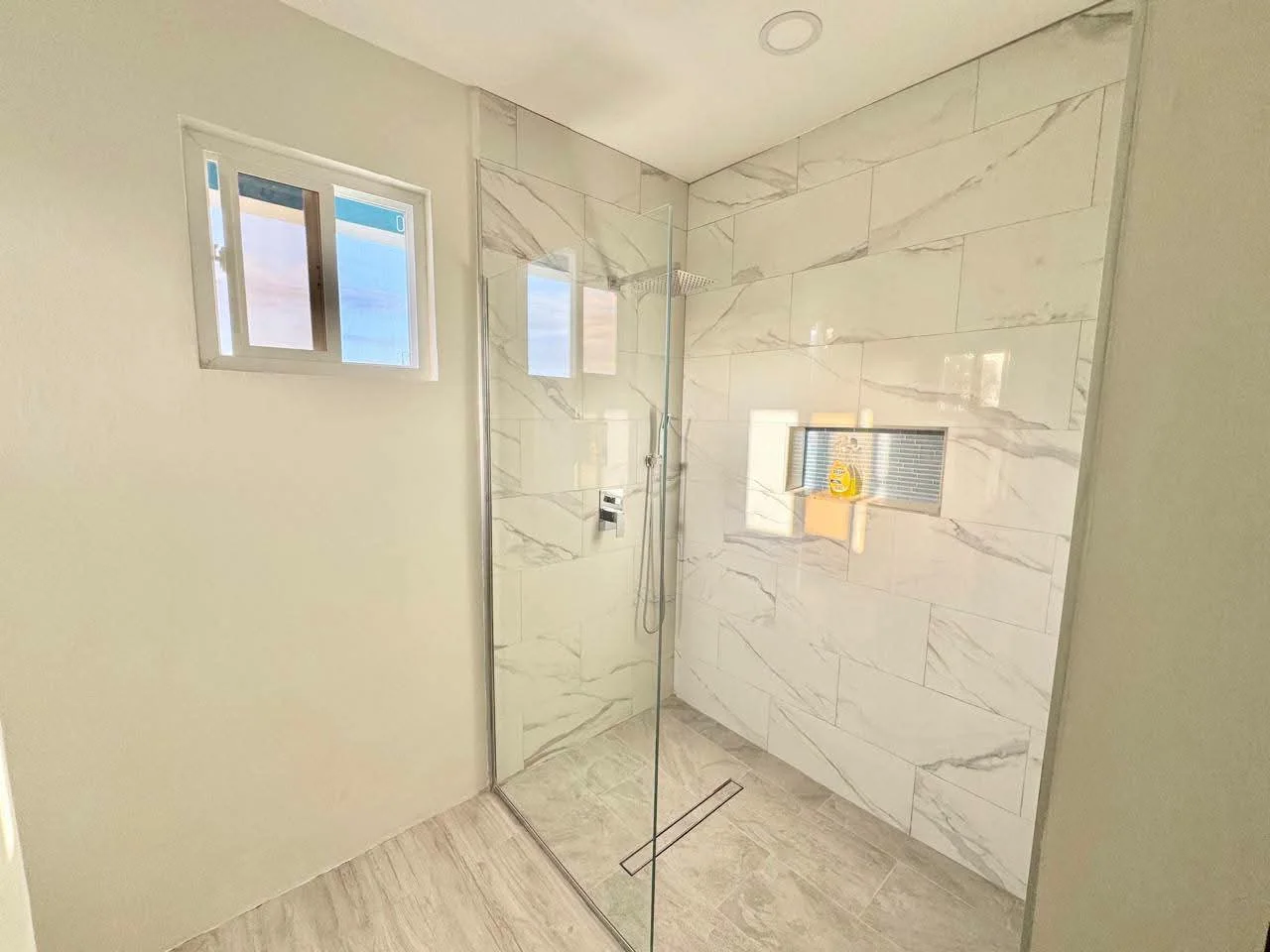 Second angle of walk-in glass shower enclosure showing marble-design porcelain wall tiles floor to ceiling, rain shower head, built-in niche shelf and linear floor drain, Saint Lucia