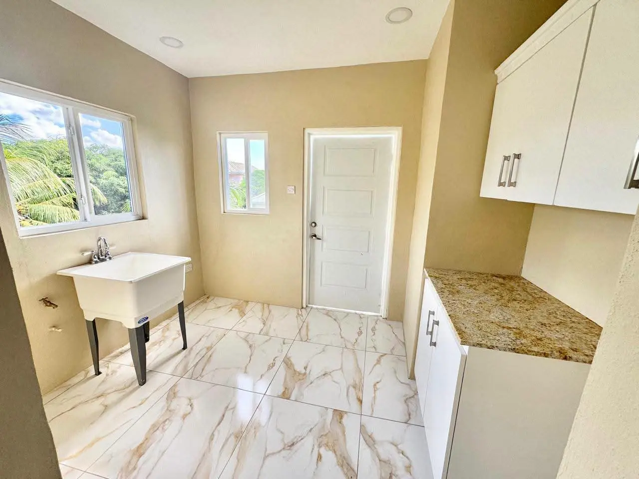 Dedicated laundry room with cream walls, marble-style porcelain floor tiles, utility sink, granite countertop, white upper and lower storage cabinets and tropical garden view through windows, Saint Lucia