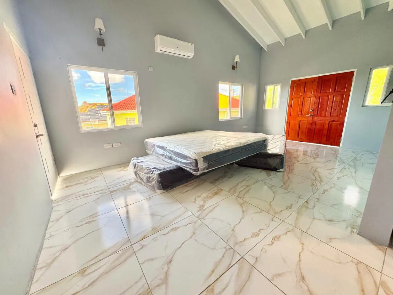 Spacious master bedroom with vaulted ceiling and exposed beams, light grey walls, wall-mounted split air conditioning unit, multiple louvre windows and king-sized bed on polished marble-design tile floor, Saint Lucia