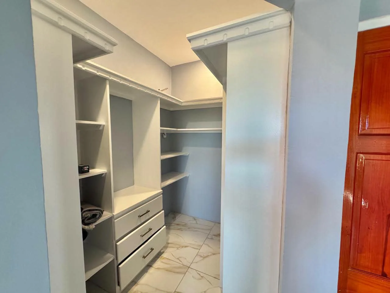 Walk-in closet in master bedroom with custom white built-in shelving, hanging rails, three-drawer chest unit and marble-design tile flooring with frosted sliding door panel, Saint Lucia